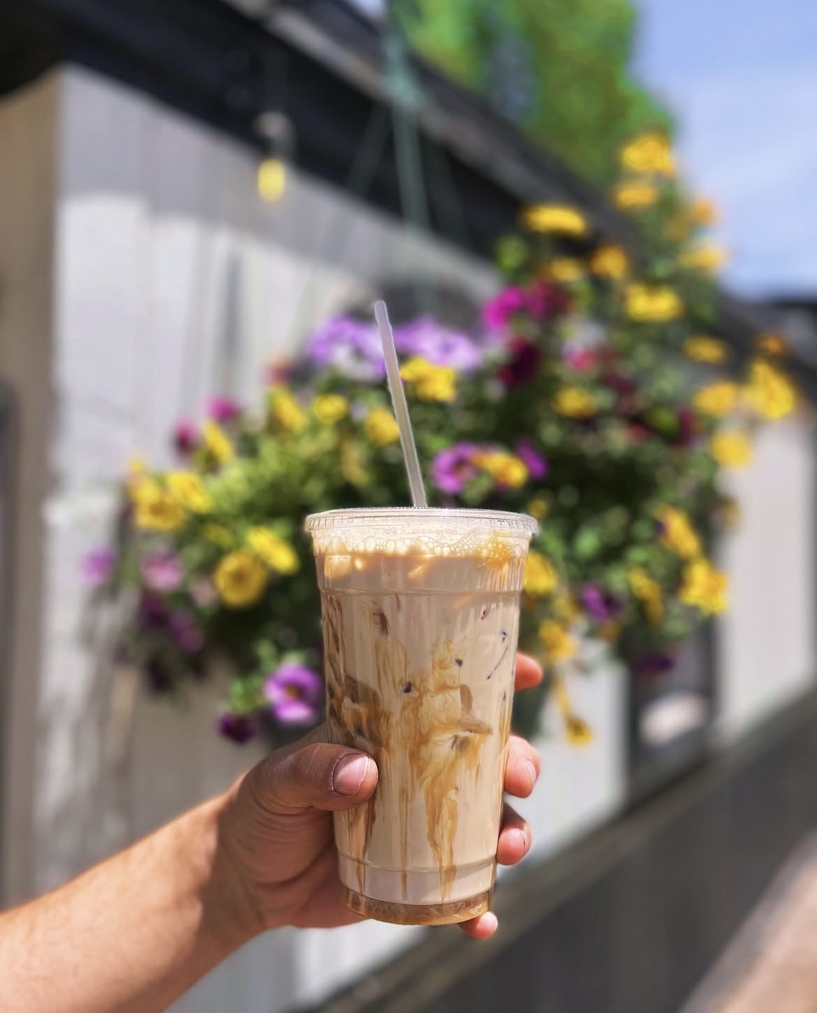 An iced coffee held up in front of hanging flower baskets outside the shop