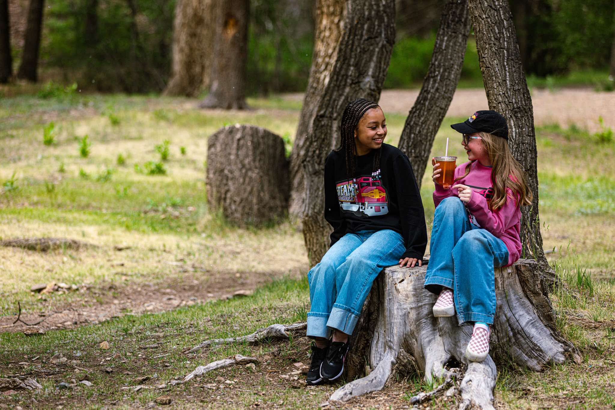 Two kids sharing an iced tea and laughing on a tree stump near the shop