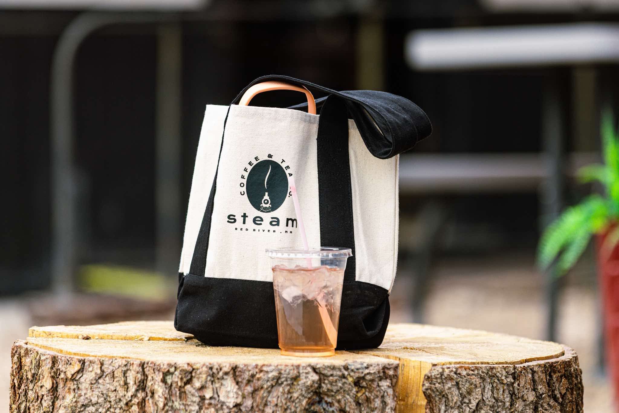 Steam Coffee tote bag and an iced tea sitting on a tree stump table